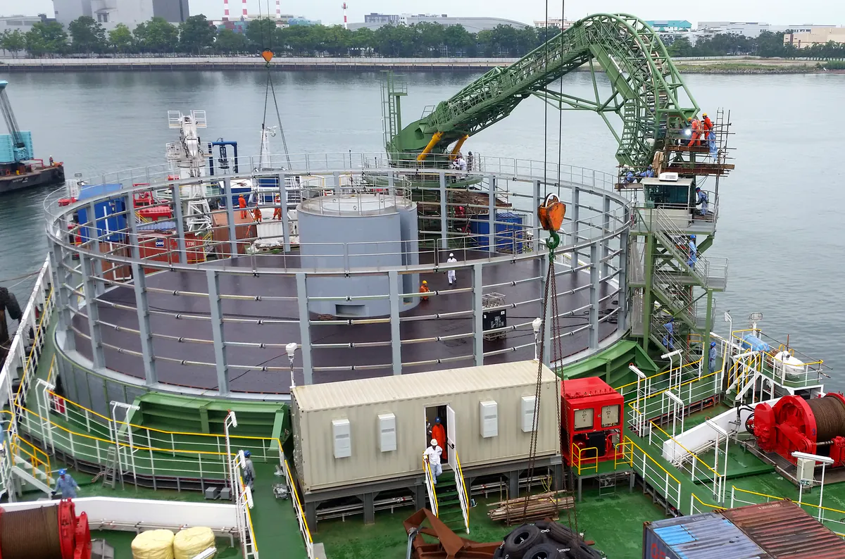 Le pont du navire câblier Ulisse&nbsp;: le carrousel circulaire qui contient le câble, la grue de guidage verte en arc et la cabine de commande.
