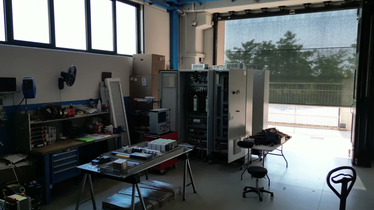 Officina D&D di Mesero: il banco di lavoro con gli utensili, due quadri elettrici aperti, una piastra di fondo appoggiata sui cavalletti e il portone aperto sui campi.